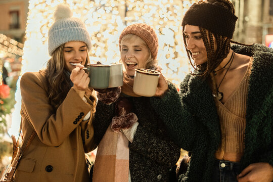 Three diverse friends drinking hot chocolate at a Christmas market at night. Happy multicultural women toasting with mugs of mulled wine, enjoying the festive holiday season. - Powered by Adobe