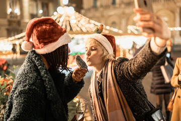 Two diverse friends taking a selfie with pet lovebirds at a Christmas market. Happy women with...