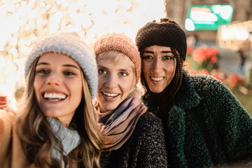 Three diverse friends taking a selfie at a Christmas market with festive lights. Happy...