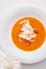 A vibrant pumpkin cream soup with shrimp and a delicate garnish of fresh white vegetable ribbons, served in a white bowl.