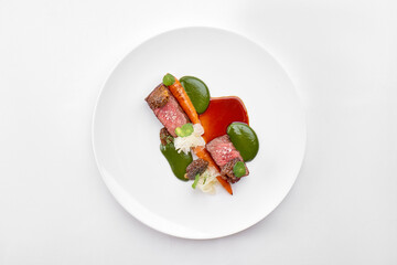 A beautifully plated steak dish with two pieces of medium-rare meat, roasted carrots, and two different sauces, served on a round white plate.