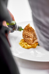 A chef's hands are drizzling a green sauce over a dish of yellow purée with a large, crispy...