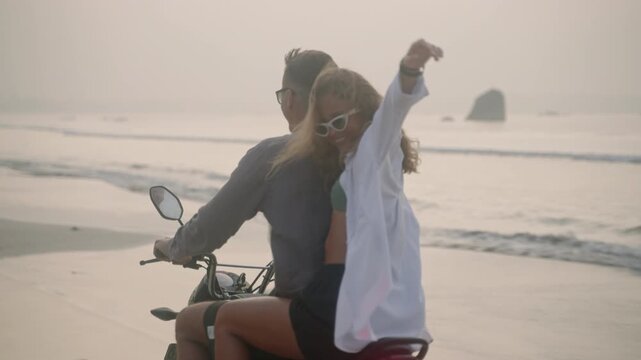 Young couple riding motorcycle on ocean beach at sunset. Man, woman travelling by motor bike on coastline along sea. Lady looks back to camera, laughs, spreads arms, touches hair blowing in the wind. - Powered by Adobe