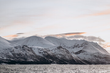 Scenic wide shot of a snow-covered mountain range reflected in a calm fjord, with soft light from a...