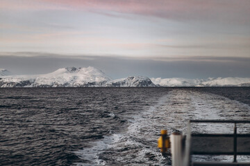A view from the back of a boat, showing the wake trailing in the dark sea. In the distance, snow-covered mountains stand under a soft, cloudy sky at dusk.