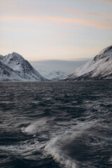 A dramatic winter seascape with turbulent, dark waves in the foreground, and a large range of...