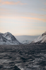A dramatic winter seascape with turbulent, dark waves in the foreground, and a large range of...