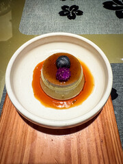 Japanese caramel pudding topped with a blueberry and purple edible flower, served on a ceramic plate with rich caramel sauce.