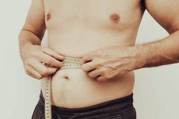A man stands in a casual setting, measuring his waist with a tape measure. Weight loss, motivation, fat burning. Overweight man measuring waist with measure tape, close up image.