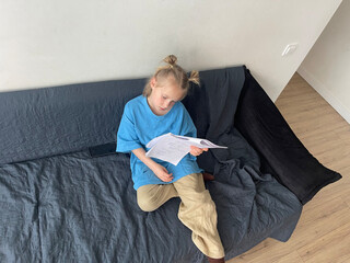 Lovely little smart child girl looking at book while sitting on sofa indoor
