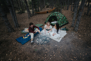 Three friends sit around a campfire in a forest during their camping trip, surrounded by a tent and...
