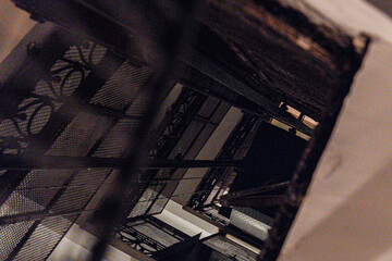 View up an old building’s interior shaft with metal grates and decorative patterns. Dimly lit...