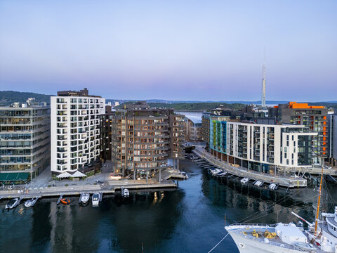Aerial view of modern buildings reflecting the twilight sky, contrasted by the dark waters of the harbor and docked boats near Aker Brygge, Oslo, Oslo, Norway.