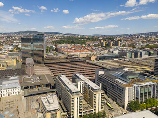 Aerial view of modern buildings casting shadows under a bright sky, contrasting with the red-tiled roofs of older structures and the distant green hills, Oslo, Oslo, Norway.