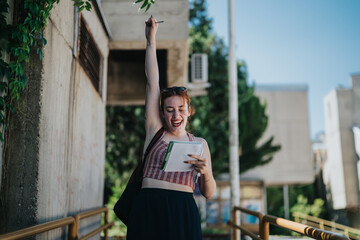 A cheerful student raises her arm in celebration, holding a notebook, outdoors on a sunny day.