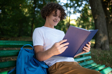 Curly-haired teenager sitting on the bench in the park and reading a book