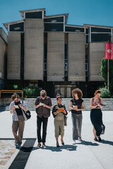 A diverse group of students and their professor walking in front of a university building, carrying devices and notebooks.