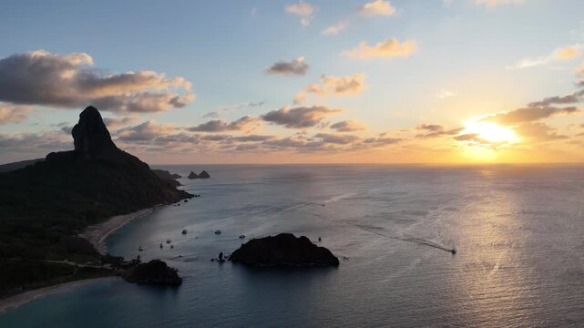 P&ocirc;r do sol sobre Fernando de Noronha: c&eacute;u em tons laranja e p&uacute;rpura refletidos no mar calmo, com silhuetas de ilhas e barcos compondo uma cena dourada e serena