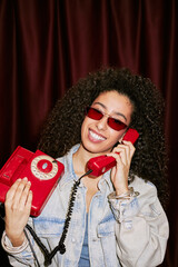 Portrait of young adult biracial woman smiling while holding vintage red rotary phone to ear in...