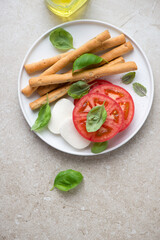 Plate with salted breadsticks and ingredients for caprese, vertical shot on a beige stone background, top view