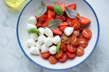 Caprese with fresh strawberry, cherry tomatoes, mini mozzarella balls and fresh basil served in a white and blue plate, middle closeup, horizontal shot