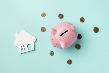 A pink piggy bank surrounded by coins with a house model on light blue background. Top view, flat...
