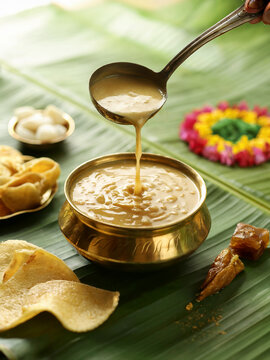 Brass uruli of creamy palada payasam with roasted cashews, on a banana leaf, ladle pouring in, surrounded by Onam snacks and flower rangoli.