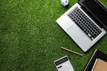 A laptop, notebook with pencil, calculator and crumpled paper on a green grassy lawn, describing...