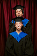 Portrait of young adult Black man and young adult biracial woman wearing graduation caps and gowns...