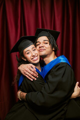 Portrait of young adult multiethnic man and woman wearing graduation caps and gowns embracing and...