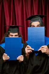 Portrait of two young adult multiethnic graduates wearing caps and gowns holding blue diploma...