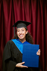 Portrait of young adult Hispanic woman smiling and holding diploma folder while wearing graduation...