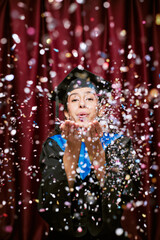 Portrait of young adult Hispanic woman wearing graduation cap and gown blowing confetti toward...