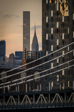 View of a bridge's intricate cable patterns framing the Chrysler Building and modern skyscrapers against a soft sunset glow, New York, New York, United States.