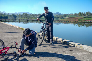 One Man Ties His Shoelace while Another Stands with His Bike Near A Calm Lake