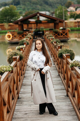The woman stands on a wooden bridge that leads to a gazebo on the water. She is dressed in a light...