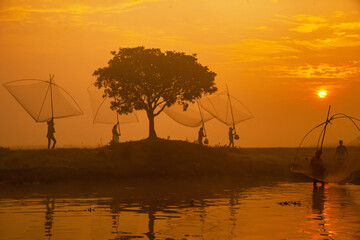 Pabna, Bangladesh - 28 November 2023: View of silhouettes of fishermen with fishing gear against the golden sunset, reflected on the tranquil river, creating a serene rural scene.