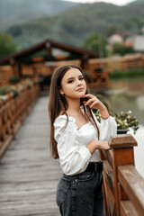 A woman stands on a wooden bridge near a pond. She is wearing a white blouse with long sleeves and...