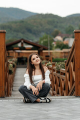 A woman is sitting on a wooden bridge by a pond. She is wearing a white blouse with long sleeves...
