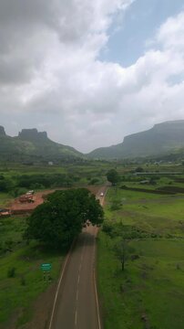 A drone view captures a car driving along the Nashik&ndash;Trimbak highway, surrounded by lush greenery and distant mountains. The dramatic cloudy sky, adds a serene charm 4K
