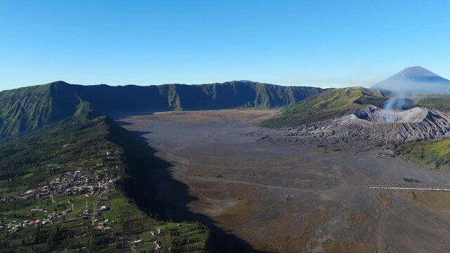 Left orbit drone shot sweeps over the Tengger caldera, revealing steaming Mount Bromo, distant Mount Semeru, and Cemoro Lawang village along the rim under a clear morning sky.