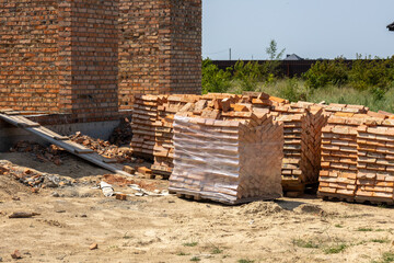 Red bricks are stacked on the ground near a brick house under construction. One brick stack is...