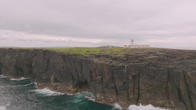 Aerial view of the stark white Eshaness Lighthouse sits atop rugged cliffs, contrasting with the turbulent sea below, Eshaness, Scotland, United Kingdom.