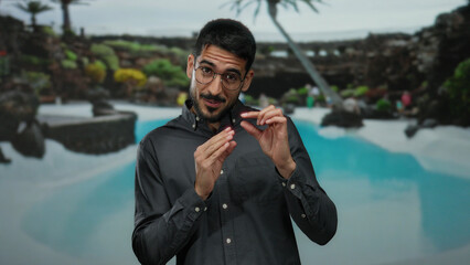 Hispanic man gestures in a serene outdoor pool setting at a luxury resort, surrounded by lush greenery and tropical ambiance.