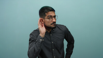 Young man listens wearing glasses on blue background, showing attentive posture with hand beside ear, expressing curiosity and focus in a portrait setting.
