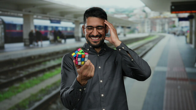 Young man smiling while holding a colorful puzzle cube at a train station, showcasing joy and engagement in a lively outdoor railway environment.