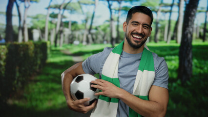 Naklejka premium Young man smiling with soccer ball in outdoor park wearing scarf vibrant greenery sunny day attractive confident lifestyle mood