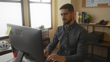 Man working on a computer in an office setting with shelves and books in the background showcasing a modern workplace environment.