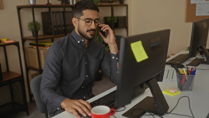 Young hispanic man working in a modern office, speaking on phone with a computer in front,...
