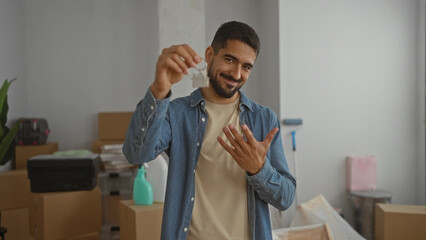 Young man holding house keys smiling in a new home interior surrounded by boxes indicating moving...